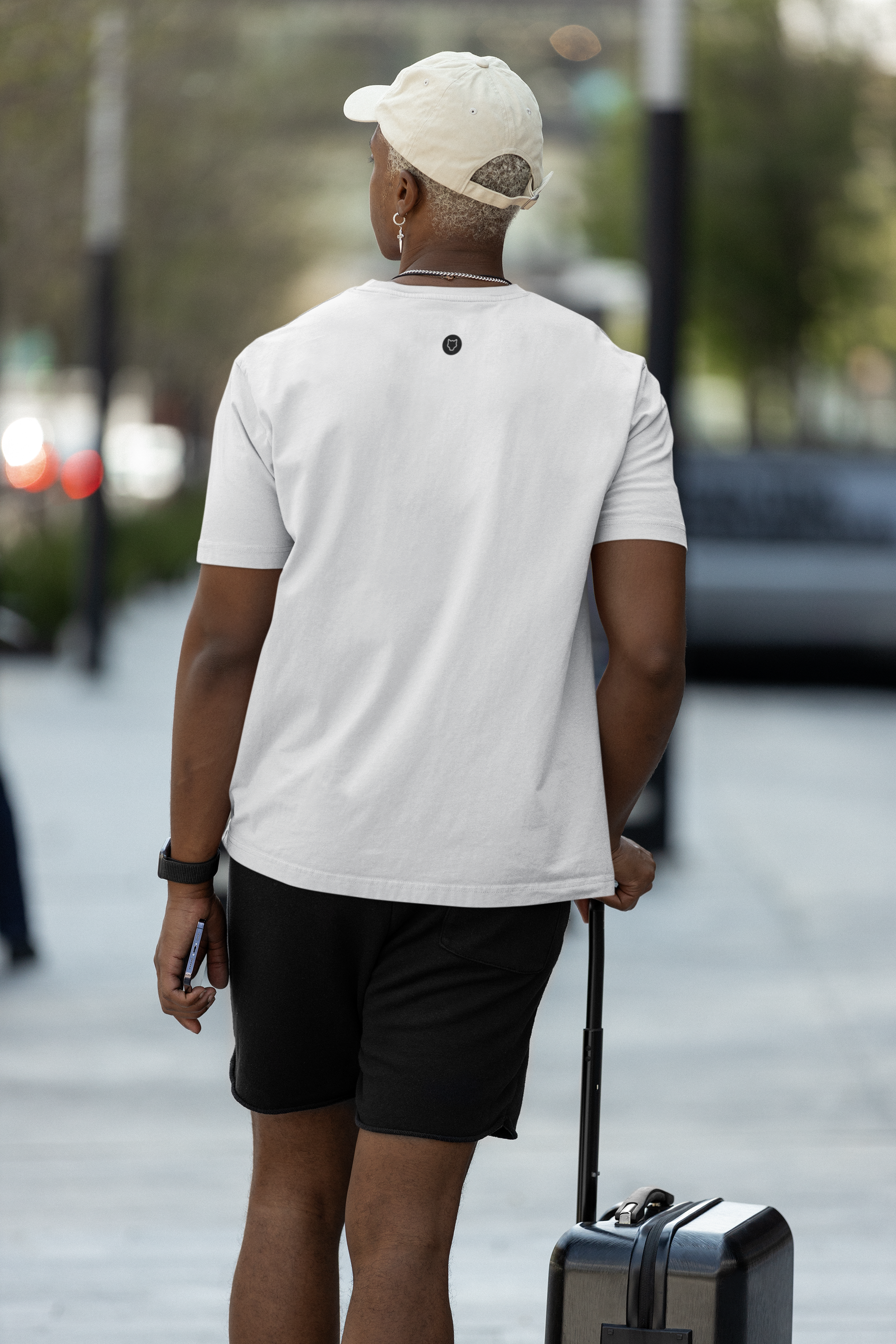 A Good looking man posing backwards wearing a white Casual T-Shirt with a suitcase in his hand. He is pairing the T-Shirt with a pair of black Shorts.