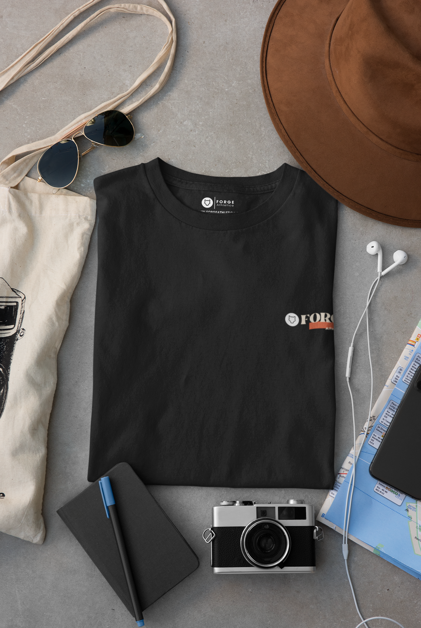 A back Unisex Round Neck T-Shirt folded perfectly and placed on a grey desk with a brown hat, a camera, a white tote bag, a diary and a pair of earphones.