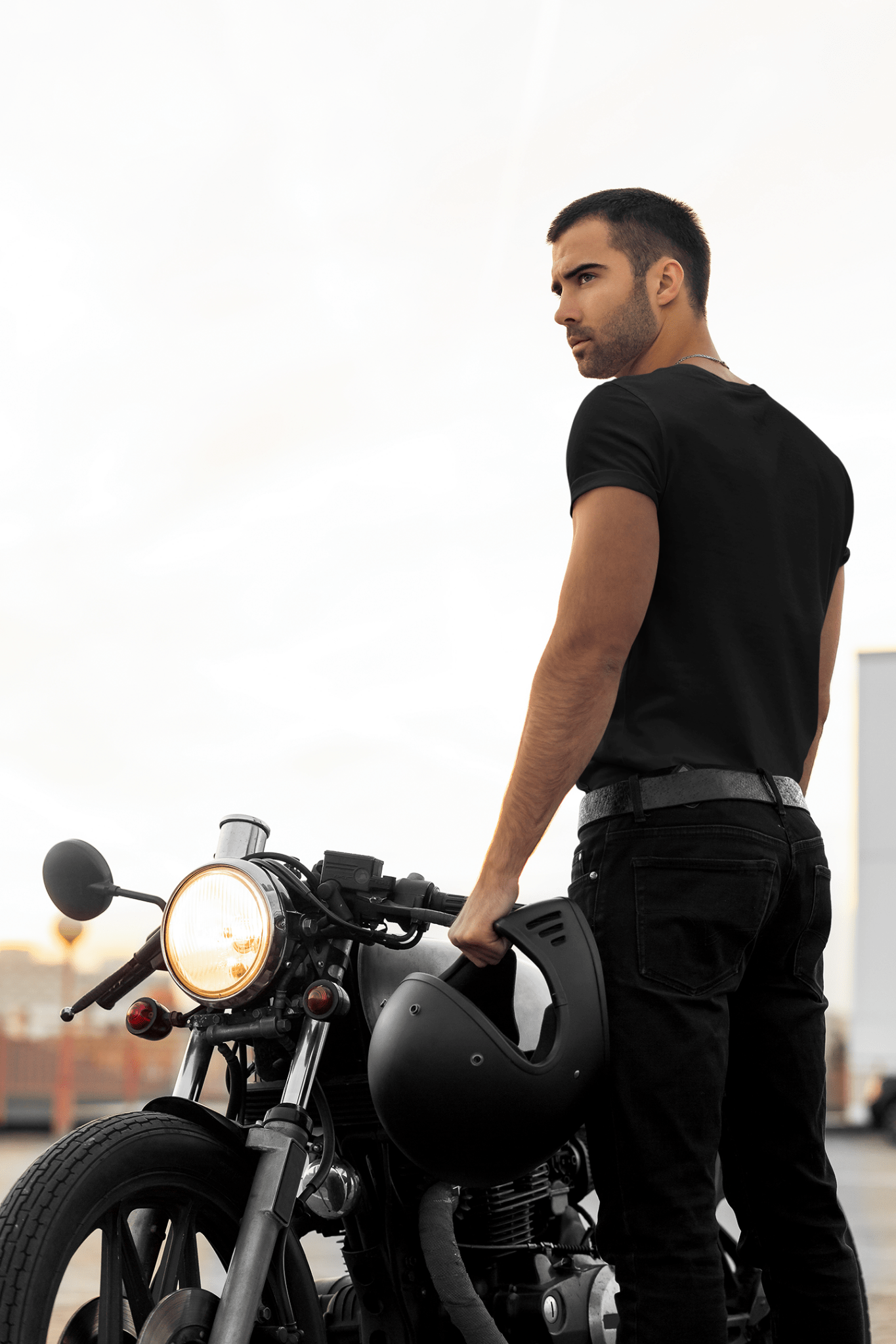 Man posing backwards wearing a black vibrant round neck T-Shirt in front of a cruiser motorcycle with a leather belt and a helmet in his hand.