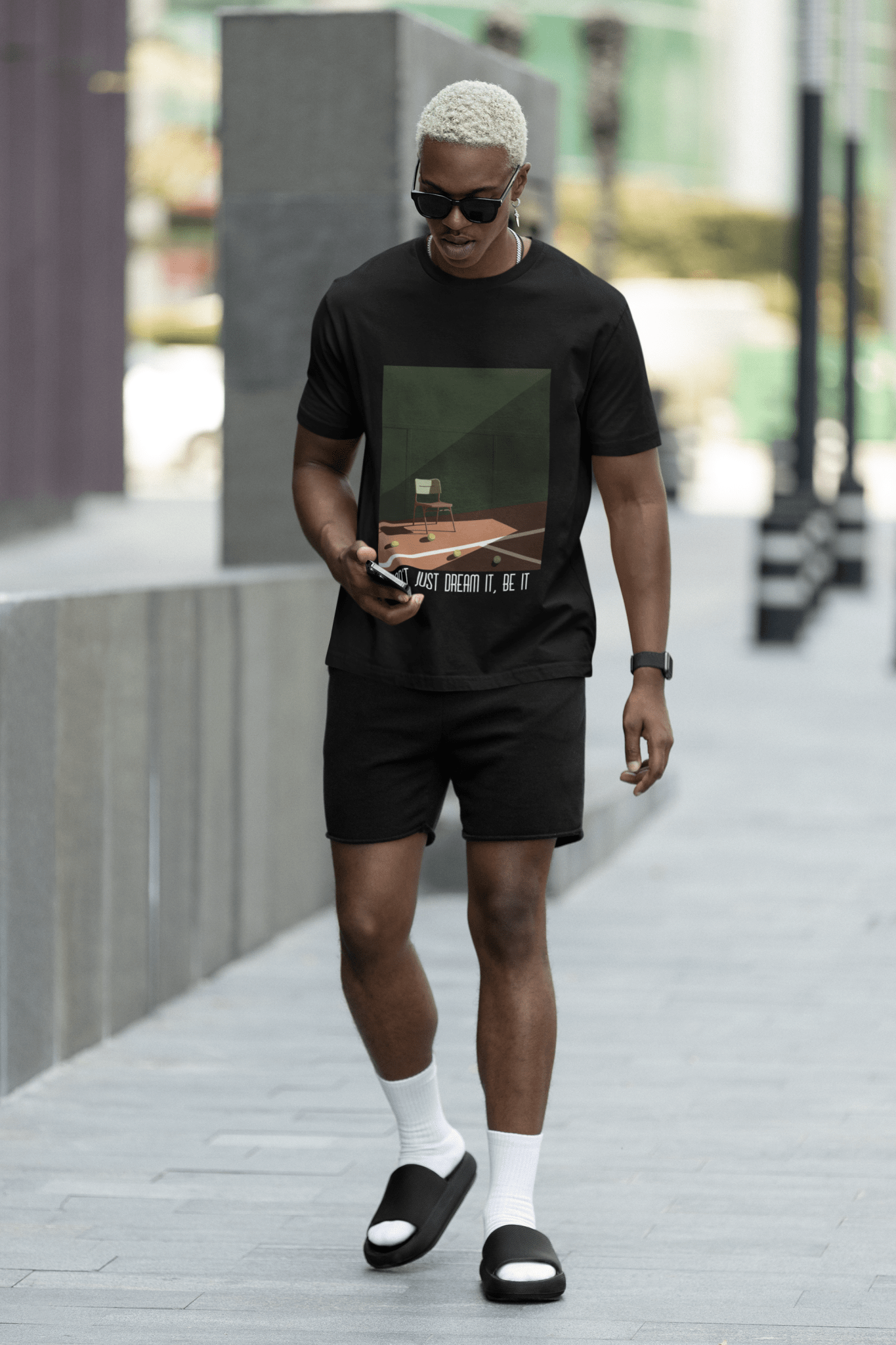 Black man wearing a black casual T-Shirt from Forge Athletica on shorts casually walking on a street using his phone. He is wearing a pair of black glasses and a pair of black sliders with white socks.